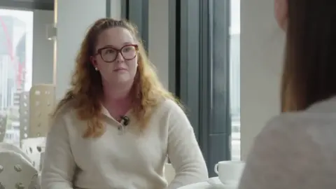 A woman in a white jumper with glasses and curly light hair sits at a table talking with the reporter.
