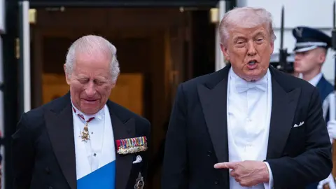 Donald Trump points his finger at a laughing King Charles as they stand side by side before entering a State Dinner at Washington DC. The two are wearing tailcoats and white shirts, while King Charles also has a blue sash.