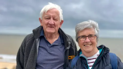 Martin Giles/BBC Malcolm and Pamela Cousins are standing on the promenade and have the sea in the background. It is a cloudy day and they are both wearing warm coats. Pamela's is blue puffer coat and Malcolm a grey fleece. They are both smiling at the camera.