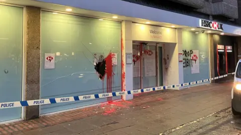 A branch of HSBC bank with red paint splashed on the glass and doors. There is a large hole in one of the window panes and lettering "DROP EIBIT" painted on a pane in black paint.