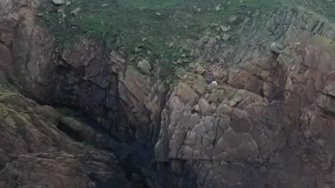 A large cliff face, the dog and the volunteer can be seen very small on the side of the cliff