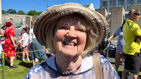 Rhoda Baxter is wearing a brown and weight hat, she has a strap over her left shoulder. She is wearing a beaded necklace and a blue and white shirt. Behind her is a crowd of people and a number of large stone pieces.