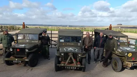 Five men posing next to three 1940s Jeeps. The vehicles have canvas roofs and are painted army green with a white star in a circle - US Army logos - on the bonnet.