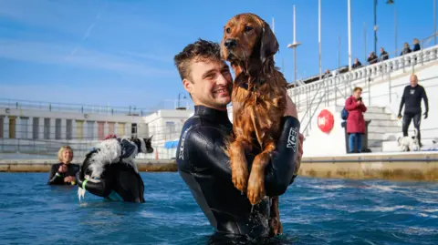 Penzance Council A golden-coloured dog being held by a man in an outdoor swimming pool. The man is wearing a wetsuit and is smiling.