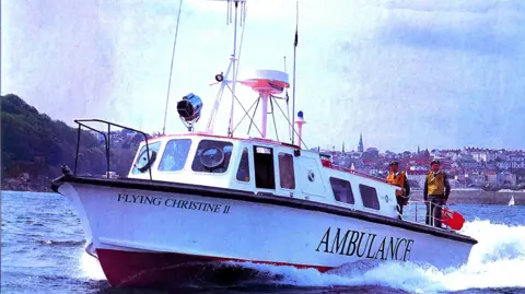 St John Ambulance & Rescue A large sailing boat used as a marine ambulance taken in the 90s in the ocean. Two men are standing on the back of the boat with life vests on. Guernsey landscape can be seen behind.