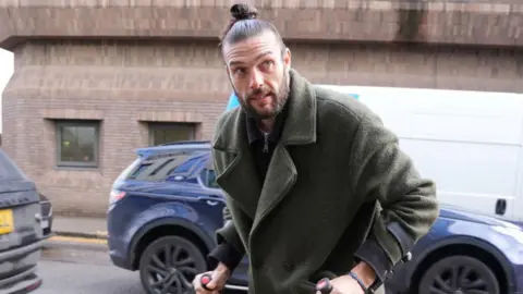PA Media Andy Carroll has his brown hair tightly tied in a bun. He has a beard and is wearing a fluffy green jacket over a zipped shirt. He grips onto black crutches with both hands.