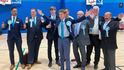 BBC Reform UK candidates in the election count venue in Coalville, Leicestershire. They all sport blue rosettes and wear suits over white shirts and light blue ties.