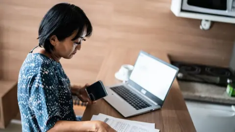 Stock photo shows a person looking down at bills and a laptop while holding a phone in their hand next to their kitchen.