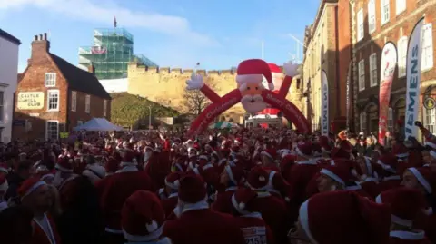 A wide shot of hundreds of people dressed in Father Christmas outfits gathering for a fun run. There is an inflatable Santa arch. 