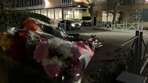 A pile of flowers stay on the wall outside the school, where the police cars and ambulances are still outside the building