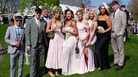 PA Media A group of men in grey suits and women in pink dresses and fascinators pose for a photo on a grassy slope