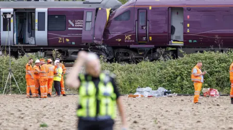 EPA/Shutterstock A police officer scratching his head is in the blurred foreground, with a backdrop of two purple train carriages. They have collided head-on. Men in orange hi-vis stand in a group on a field beside the track.
