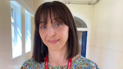 Dr Anderson, who has long dark hair with a fringe, is in a narrow corridor with high ceilings. It looks like a very old building with magnolia painted walls and a red floor. Behind her is a blue door which leads further down the corridor, and a small window. She is wearing a bright coloured patterned top, and a red lanyard which hangs around her neck.