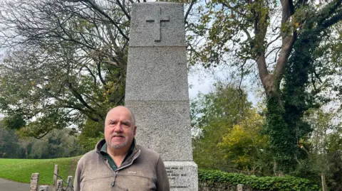 Steve Davies wearing a brown jumper over a green shirt. He is smiling at the camera and is stood in front of the grey war memorial. Behind the memorial is trees and grass. 