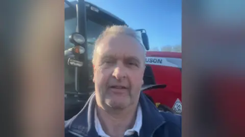Jeremy Padfield A man taking a selfie in front of a red tractor. He is wearing a blue jumper and the sky is blue in the background.