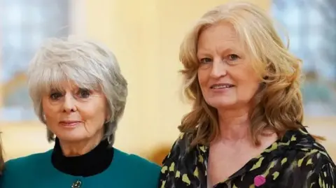 PA Media Head and shoulders image of Diana Parkes and Hetti Barkworth-Nanton, standing side by side and looking at the camera. Diana has grey, short hair and is wearing a jade coloured suit jacket with a black roll neck jumper. Hetti has dark blond, shoulder length, wavy hair and is wearing a black and yellow shirt.
