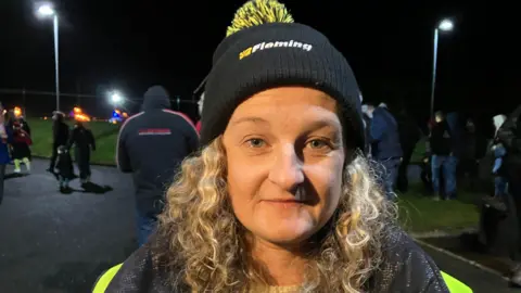 Catherine McCombe standing outside in the dark, she has curly long blonde hair and is wearing a black and yellow hat with a high vis jacket on.