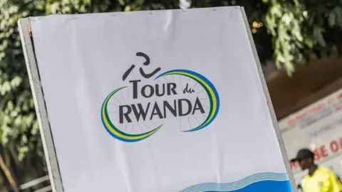 A white and blue banner with the logo of cycling event Tour du Rwanda on it, which is a pictogram of a figure riding a bike with the event name contained within the two wheels, with those wheels made up of the colours of the Rwandese flag.