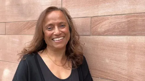 BBC/Marian McNamee Malkit has light brown hair and is wearing a black top with a gold necklace. She is smiling and standing in front of a red sandstone wall.