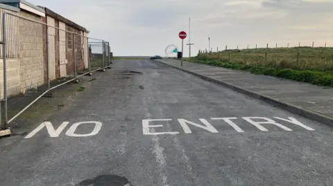 LDRS 'NO ENTRY' written on the road in white letters. There is also a red stop sign further down the road. To the left there is a bordered up brick building with metal fences in front of it.