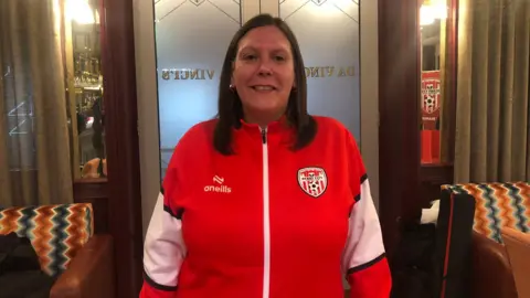 Derry City Supporters’ liaison officer Karen Pyne is wearing a red and white club tracksuit and is standing at the door of a public house.