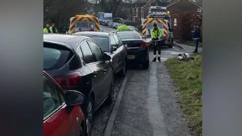 Four cars parked on a road with a tow-away truck loaded with cars ahead of them. A man in high-vis jacket is making notes. Three other men in high-vis jackets can be seen nearby with another tow truck. 