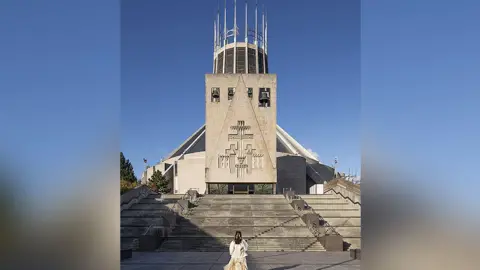 Historic England Exterior view of the Metropolitan Cathedral of Christ the King, Mount Pleasant, Liverpool. It shows the back of a female wearing a cream jacket and dress with long brown hair approaching the bottom of the steps to the entrance.