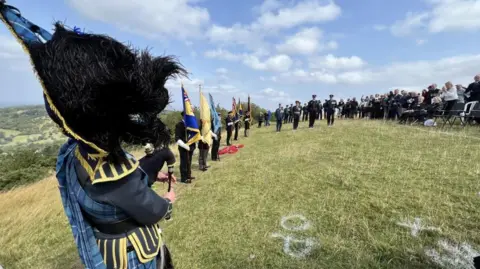 BBC A group of people watching a military service on on Cleeve Hill, including several people in military uniforms and a bagpiper, to remember seven men who died 