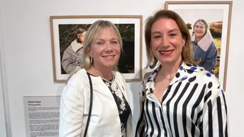 Two white women in white shirts stand smiling in front of photos at an exhibition. 