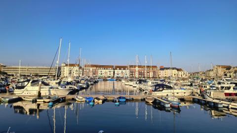 Weymouth seafront as seen from the harbour, with several boats docked in front of the buildings that overlook the water