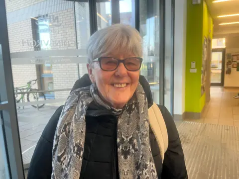A woman with grey hair, glasses and a black and white scarf smiles in the corridor of a medical centre. 