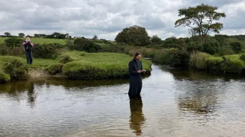 Reverend Robin Thwaites in the river on Bodmin Moor, wearing robes and holding a book, while a man with a white beard overlooks from the far, grassy bank