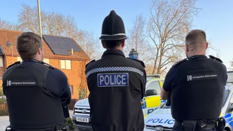 A police officer stands in the middle of two immigration enforcement officers who all have their backs turned to the camera. Two police cars are parked in front of them.