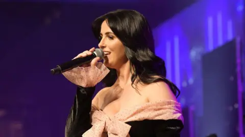 Lisa McHugh is singing into a black microphone. She is wearing a black dress with a nude poke a dot bow at the front. She has long black hair. The background is various shades of blue and purple. 