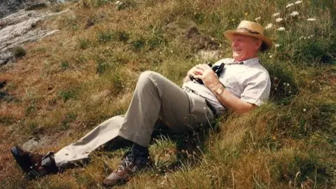 Dougie's Grant Douglas Le Huquet laying on a grass bank in Guernsey. He is wearing a straw hat and holding binoculars.