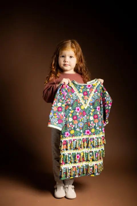 Kieran Dodds Full-length studio portrait of a young child with long curly hair standing against a dark brown background and holding up a colourful floral-patterned dress. The dress features bright flowers, mushrooms, and decorative tassel trim. The child wears a long-sleeved brown top, grey trousers, and light-coloured shoes. Warm lighting highlights the vibrant colours of the garment.