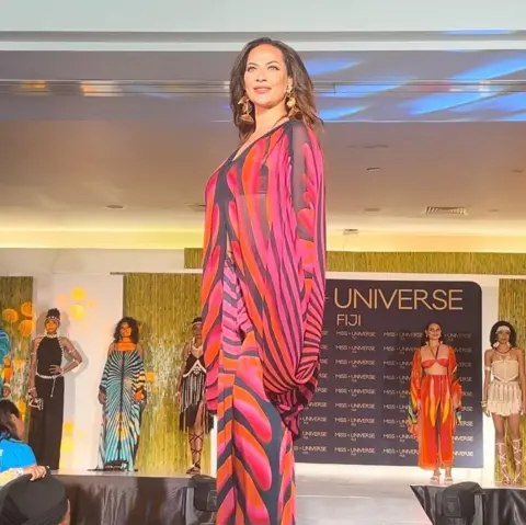 JENNIFER CHAN A woman in the foreground wearing a red, pink, black and orange outfit poses on stage. Behind her in the background are her seven fellow contestants for the Miss Fiji contest.