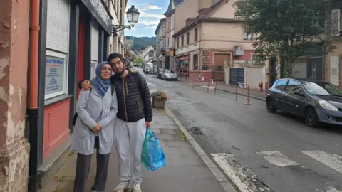 Family Missing Ali Durrani with his mother Mahjabeen. The pair are on an old-town looking street and posing for a photo. She is wearing a headscarf and a long cardigan. He has on a black outdoor jacket.
