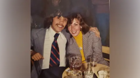 Family handout A young man wearing a suit, smiles at the camera, as he hugs his wife, who is also smiling at the camera, wearing a yellow top and checked blazer as they sit in a pub.