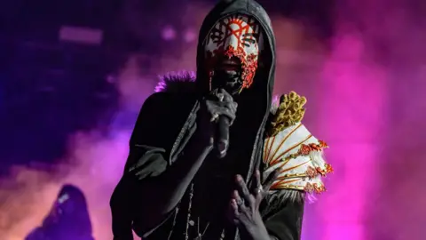 Getty Images The lead singer of Sleep Token sings a note on-stage as pink steam - illuminated by coloured stage lights - swirls around behind him. He's wearing a black, hooded cape with an ornate white and gold shoulder decoration on his left side. Under his hood, a white mask, painted with a red, geometric design, is visible. His mouth is uncovered by the mask, highlighted by jagged red edges.