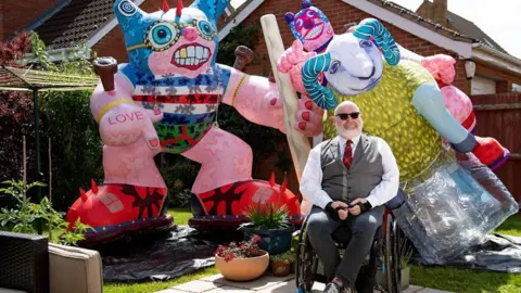 Jason Wilsher-Mills with short white hair and beard wearing sunglasses, a grey waistcoat, dark trousers, white shirt and red tartan ties sits in a wheelchair in front of two large works of art which are colourful statues of a ram and a cat like figure standing on its back legs