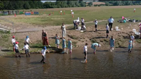 Jamie Niblock/BBC A group of children stand in a river wearing their school uniforms while carrying out water sampling using nets and trays