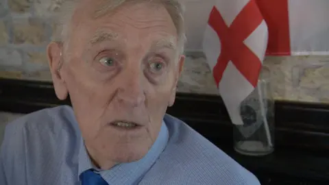 Eric Woolfenden in a pub with a glass with an England flag inside on a shelf behind him