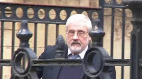 BBC Stephen Cox, wearing a buttoned-up shirt and tie, but with the top button undone, and a purple jumper and dark jacket, walking out of Reading Crown Court, with the bars of railings in front of him as he walks down a ramp outside the building