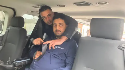 Ahsan is sitting in his wheelchair inside his specially adapted mobility car. His brother Mohsin is attaching Ahsan's seatbelt. 