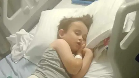 Supplied A young boy wearing a grey vest sleeping on a hospital bed. 