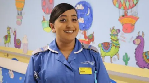 A woman wearing a nurses uniform stands in front of a colourful wall.