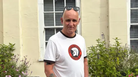 A man stands in front of two sash windows. Flower pots are in the foreground. He is in a white T-shirt and khaki shorts, with sunglasses on his head.
