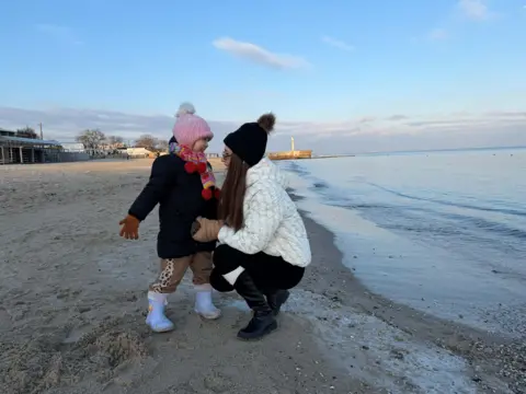 A woman and a toddler wearing warm tops and hats embrace on the beach