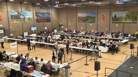 Votes are counted in a large hall. Several rows of tables are laid out with volunteers counting ballots. A number of banners advertising Northumberland decorate the walls.